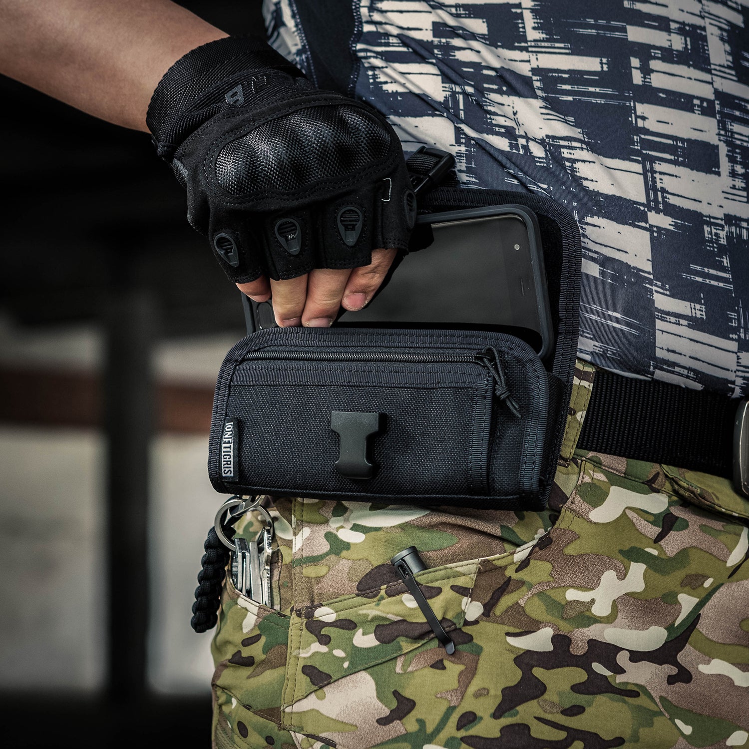 Person wearing tactical gloves and holding a black tactical pouch with camouflage pants.